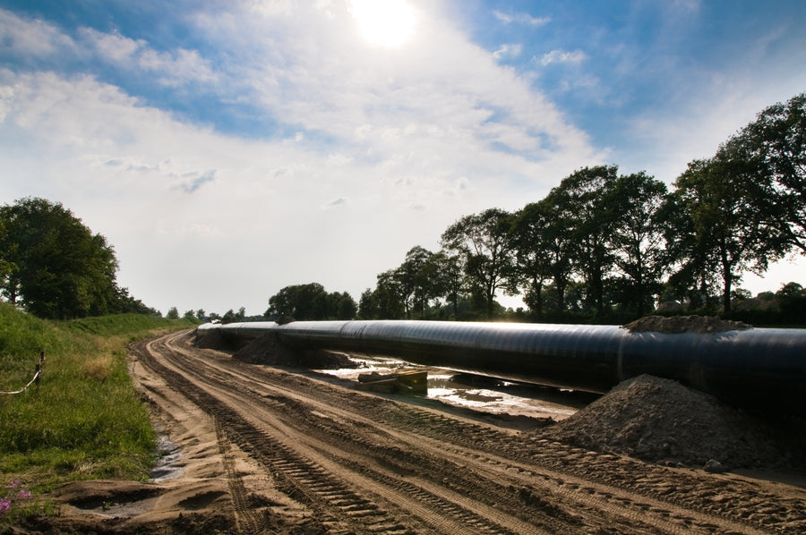 A large black industrial pipeline running along a dirt road outdoors, with trees, grass, a dirt pile, and a blue sky with clouds in the background.