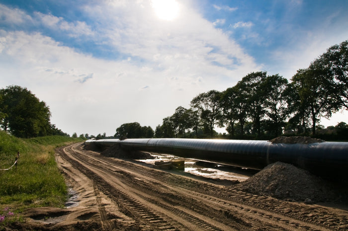 A large black industrial pipeline running along a dirt road outdoors, with trees, grass, a dirt pile, and a blue sky with clouds in the background.